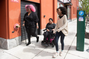 Three Black and disabled folx cracking up while strolling down a sidewalk on a windy day. On the left, a non-binary person walks with a cane in one hand and a tangle stim toy in the other. In the middle, a non-binary person rolls along in their power wheelchair. On the right, a woman is walking with fabulously windswept hair. A street parking meter is in the background on the right.