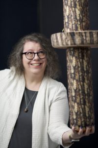 The headshot of a light-skinned, middle-aged woman with brown and grey curly hair and round, dark-blue glasses. She balances a stack of fake logs in one hand, to our right, and smiles and looks warily up at them. She is dressed in a pale ivory blazer over a dark grey shell blouse and wears a wood and silver acorn pendant necklace.
