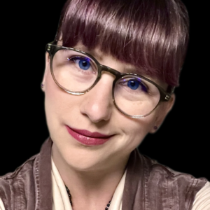 The headshot of a light-skinned woman with green-framed glasses. They smile slightly at us with their head tilted to our right. Rose gold dyed bangs cover their forehead, complementing their dusty pink vest over a light tan blouse.